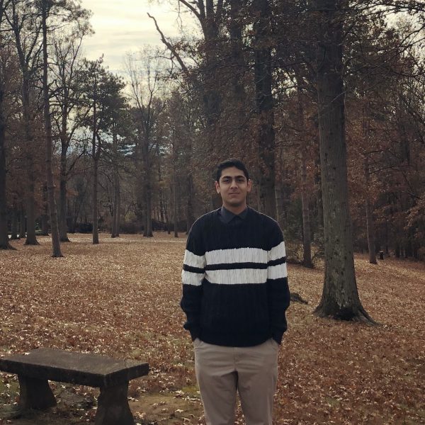 Aksir Bhura standing in front of a wooded area and beside a wooden bench wearing a black sweater with two parallel white stripes and khaki pants